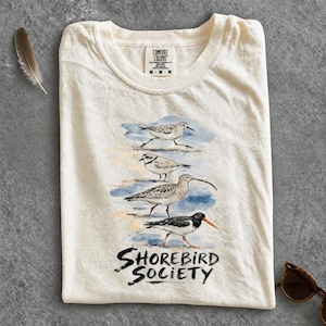 Op de afbeelding: Een crèmekleurig T-shirt met een aquarelillustratie van vier verschillende kustvogels die verticaal zijn gerangschikt. Onder de vogels staat de tekst "SHOREBIRD SOCIETY" in een zwart, handgeschreven lettertype. Het shirt heeft een ronde hals.