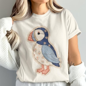 May include: A cream-coloured short-sleeve t-shirt featuring a central graphic of a puffin bird designed with a patchwork quilt aesthetic. The puffin illustration incorporates various blue, white, and orange floral and polka dot fabric patterns.