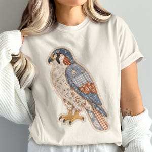 Op de afbeelding: Een crèmekleurig T-shirt met korte mouwen en een grote afbeelding van een valk. De vogel is ontworpen met een patchwork-esthetiek, met diverse patronen in tinten blauw, bruin en wit, voorzien van gele klauwen en een donkere snavel.