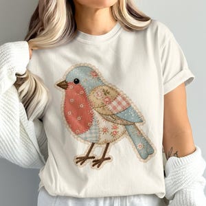 Op de afbeelding: Een crèmekleurig t-shirt met korte mouwen en een grote afbeelding van een vogel in een patchwork-stijl. De vogelillustratie bevat diverse bloemen- en ruitpatronen in tinten blauw, roze en beige met zichtbare stiksels.