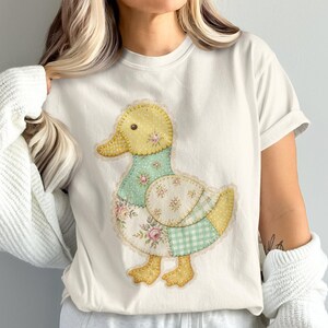 Op de afbeelding: Een crèmekleurig T-shirt met korte mouwen en een grote centrale afbeelding van een eend, samengesteld uit verschillende stoffen patronen in patchworkstijl. Het eendontwerp bevat gele stippen, lichtgroen gingham en bloemenprints in roze en wit.