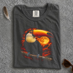 Może przedstawiać: Ciemnoszary t-shirt z nadrukiem graficznym przedstawiającym tukana z dużym pomarańczowo-żółtym dziobem siedzącego na gałęzi. Tło ukazuje żywy pomarańczowy zachód słońca z pełnią księżyca i chmurami. Metka na karku zawiera napis Comfort Colors.