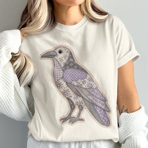 Può includere: Una t-shirt a maniche corte color crema con una grande grafica di un uccello, un corvo, disegnato con un motivo patchwork di varie texture di tessuto floreale e geometrico nei toni del viola chiaro, grigio e bianco.