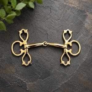 May include: A gold-coloured metal horse bit featuring a central jointed mouthpiece and two ornate, symmetrical side pieces with decorative cutouts. The hardware has a polished, reflective finish and a distinct, intricate shape on both ends.