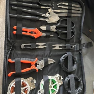 May include: A black zippered carrying case containing a set of gardening tools, including orange and black pruning shears, metal tweezers, small shovels, scissors, and spools of brown, green, and black wire secured by elastic straps.