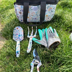 May include: A gardening set featuring a black and floral patterned tote bag, a matching hand trowel, a hand rake, and pruning shears, alongside a pair of grey and teal work gloves. The tools and bag feature a colorful floral print.