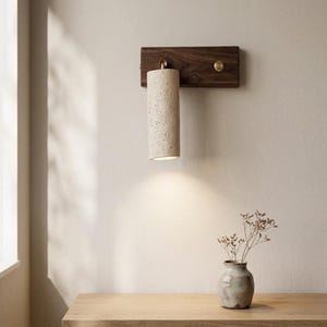 May include: A wall-mounted light fixture featuring a rectangular dark wood base with a brass accent and a cylindrical, speckled beige stone shade. The light emits a downward glow, illuminating the wall below the fixture.