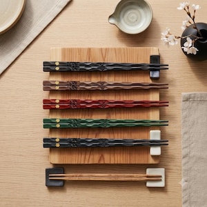 Peut inclure: Un ensemble de cinq paires de baguettes en bois texturé dans des tons noir, marron, rouge, vert et bois naturel, chacune avec un petit accent floral doré près du sommet, posées sur une planche en bois rectangulaire avec deux repose-baguettes.