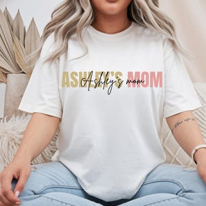Puede incluir: Una camiseta blanca de manga corta con el texto ASHLEY'S MOM en fuente de bloque con un degradado dorado y rosa, superpuesto con el texto Ashley's mom en letra cursiva negra. La camiseta tiene un cuello redondo y un ajuste relajado.