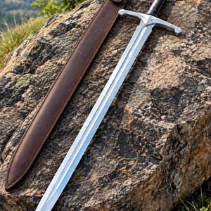 May include: A double-edged steel sword with a central fuller, a silver-toned crossguard, and a black grip topped with a silver pommel. Beside the sword lies a brown leather scabbard featuring a single strap with a metallic snap button closure.