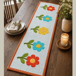 May include: A rectangular quilted table runner featuring a light blue background with a textured surface. It is decorated with five appliqued flowers in red, blue, and yellow, each with green stems and leaves, and finished with an orange border.