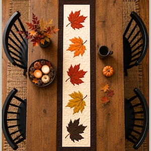 May include: A rectangular table runner featuring a cream-colored center panel with five appliqued maple leaves in shades of red, orange, yellow, and dark brown, bordered by a dark brown fabric frame with intricate quilted stitching throughout.