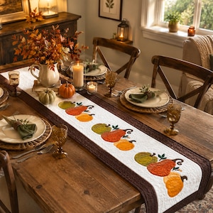 Peut inclure: Un chemin de table matelassé blanc orné d'un motif brodé de citrouilles orange, jaunes et vertes avec des tiges brunes. Le chemin de table présente des bordures texturées brun foncé et est posé sur une table à manger en bois.