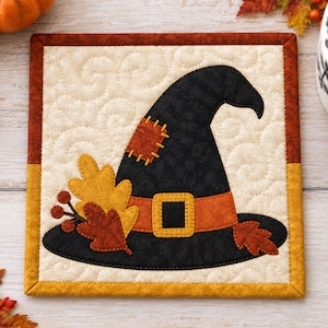 Könnte beinhalten: Ein quadratischer, gesteppter Stoffuntersetzer mit einem schwarzen Hexenhut, einem orangefarbenen Band mit Schnalle, einem gelben Flicken sowie Herbstblättern und Beeren. Der Hintergrund ist cremefarben mit Wirbelsteppung und einem orangefarbenen Rand.