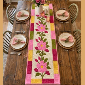 Puede incluir: Un camino de mesa acolchado rectangular con una enredadera vertical central de tres grandes flores rosas y hojas verdes, bordeado por bloques de patchwork coloridos en rosa, amarillo y verde, sobre una mesa de madera con cuatro cubiertos.