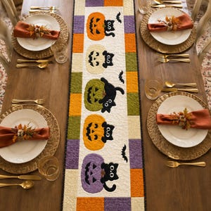 May include: A quilted table runner featuring five colorful pumpkins and black cats, accented with small black bats. The runner is bordered by a patchwork of orange, purple, and cream squares, set on a wooden dining table with four place settings.