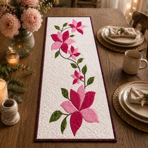 May include: A rectangular table runner with a white textured background featuring a central floral pattern of pink and magenta flowers with green leaves. The runner has a dark purple border and a quilted surface texture throughout the fabric.