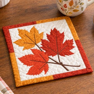 Peut inclure: Un dessous de verre carré en tissu matelassé avec un fond blanc et un motif piqué de trois feuilles d'érable dans des tons orange et rouge. Le dessous de verre présente une bordure composée de bandes de tissu orange et rouge alternées.