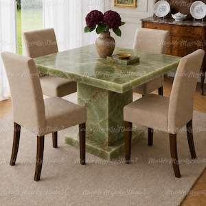 Puede incluir: Una mesa de comedor cuadrada con una superficie de ónice verde pulido y una base de pedestal a juego, acompañada de cuatro sillas tapizadas con asientos de tela beige y patas de madera marrón oscuro. Un jarrón de cerámica con flores oscuras.