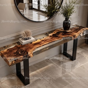 May include: A long console table featuring a live edge wood slab top with a natural grain pattern encased in clear resin. The table is supported by two black metal U-shaped legs. A stack of books and a decorative object sit on the surface.