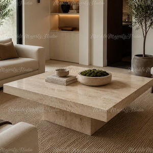 May include: A square, low-profile coffee table crafted from beige travertine stone with a natural porous texture. The table features a thick, flat tabletop supported by a central block base. A small bowl and two stacked books sit on the surface.