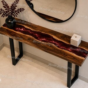 May include: A rectangular console table featuring a natural wood slab top with a central river-like inlay of deep red resin. The table is supported by two black metal U-shaped legs and is positioned against a wall with a black-framed oval mirror.