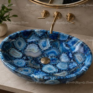 May include: An oval vessel sink crafted from polished blue agate slices with visible crystalline centers and natural banding. The basin features a smooth, glossy finish and a central gold-toned drain, set against a neutral stone countertop.