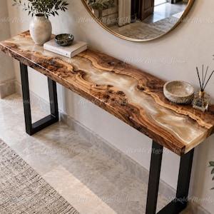 Op de afbeelding: Een rechthoekige sidetable met een houten blad met natuurlijke randen en een centrale inleg van doorschijnende witte hars. De tafel rust op twee zwarte, U-vormige metalen poten. Op het blad staan een keramische vaas, een boek en een geurstokjes.