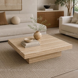 Puede incluir: Una mesa de centro cuadrada y baja fabricada en piedra travertino beige con una textura porosa natural. La mesa presenta un tablero grueso que descansa sobre una base empotrada, decorada con un cuenco pequeño, dos libros apilados y un florero.