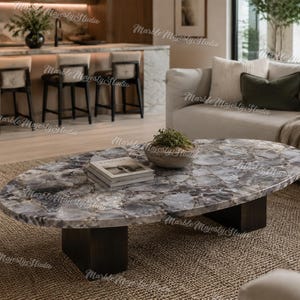 Puede incluir: Una mesa de centro ovalada con un tablero de piedra ágata gris pulida con patrones cristalinos naturales y variaciones de tono. La mesa se apoya sobre dos bases de bloques de madera maciza oscura, proporcionando una base estable.
