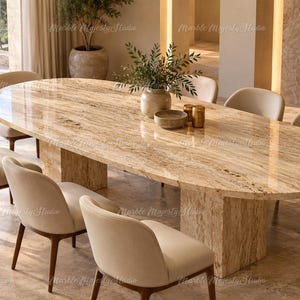 Op de afbeelding: Een ovale eettafel gemaakt van gepolijste beige travertijnsteen met een opvallend natuurlijk aderpatroon, ondersteund door twee dikke rechthoekige voetstukken, omringd door zes gestoffeerde stoelen met lichtcrème stof en houten poten.