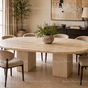 Pode incluir: Uma mesa de jantar oval com tampo de pedra travertino bege claro e duas bases retangulares correspondentes. A mesa é rodeada por seis cadeiras com pernas de madeira escura e assentos estofados em bege claro com encostos curvos.