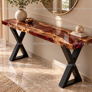 Puede incluir: Una mesa consola rectangular con un tablero de madera de borde natural y una incrustación de resina roja oscura brillante en el centro. La mesa se apoya sobre dos patas de metal negro en forma de X, proporcionando una base estable.