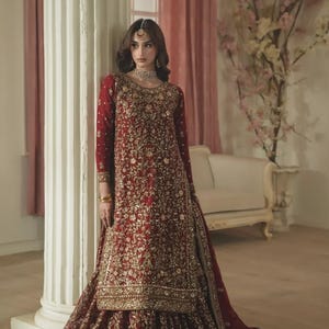 May include: A deep red long-sleeved tunic and floor-length skirt set featuring intricate gold embroidery and beadwork throughout. The ensemble includes a matching red dupatta with a decorative border, a gold choker necklace, and a gold headpiece.