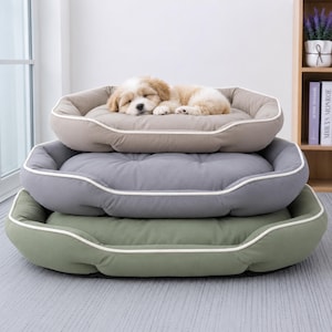 May include: A stack of three rectangular pet beds with raised edges and white piping trim. The top bed is beige with a small, light-coloured puppy sleeping on it. The middle bed is dark grey, and the bottom bed is sage green. Each bed features a tufted centre.