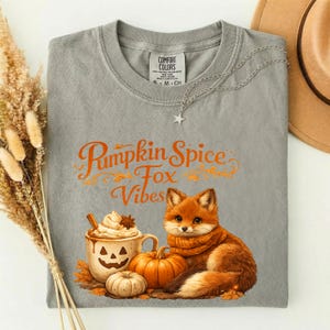 May include: A gray t-shirt featuring a graphic of a fox, a pumpkin, and a mug with a jack-o'-lantern face. The text reads "Pumpkin Spice Fox Vibes". The shirt has a crew neck and a Comfort Colors label. A silver star necklace rests on the collar.