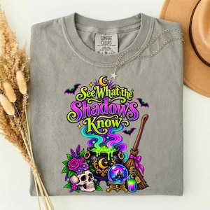 May include: A grey t-shirt featuring a graphic print with the text "See What the Shadows Know" in purple and green. The design includes a black cauldron with green liquid, a skull, purple flowers, a crystal ball, and a broomstick.