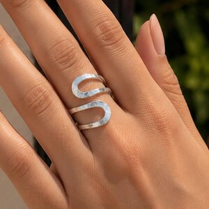 Op de afbeelding: Een zilverkleurige ring met een open bypass-ontwerp, voorzien van twee gebogen, parallelle uiteinden die om de vinger wikkelen. Het metalen oppervlak heeft een subtiele, gehamerde textuur die het licht opvangt voor een glanzende uitstraling.