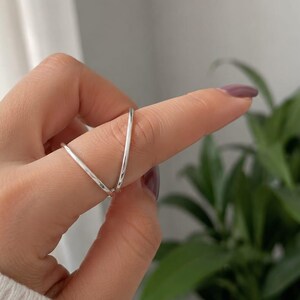 Op de afbeelding: Een zilveren ring met een V-vormig ontwerp dat om de vinger wikkelt. De band is dun en glad met een gepolijste metalen afwerking. De ring is zo geplaatst dat de open, hoekige structuur over het vingergewricht goed zichtbaar is.