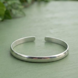 May include: A polished silver-tone cuff bracelet with a smooth, rounded surface and a simple open-ended design. The metal band features a consistent thickness throughout its circular shape, creating a clean and reflective appearance for the wrist.