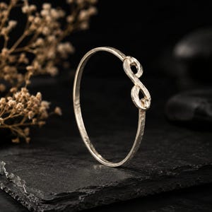 May include: A silver-toned bangle bracelet featuring a textured, hammered finish and a prominent infinity symbol design integrated into the circular band. The metal surface reflects light, highlighting the uneven, handcrafted appearance of the jewelry piece.