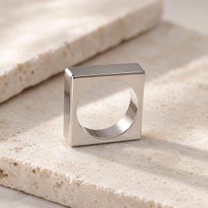 Könnte beinhalten: Ein polierter silberfarbener Ring mit einem markanten quadratischen Außenprofil und einer kreisförmigen Innenöffnung. Die Metalloberfläche ist glatt und reflektierend, mit sauberen, scharfen Kanten und einer soliden, einheitlichen Konstruktion.
