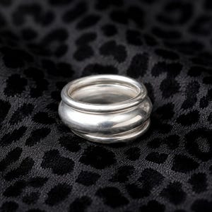 Può includere: Un anello in argento lucido caratterizzato da un design a più fasce con tre strati distinti, lisci e arrotondati, sovrapposti per formare un unico gioiello con una superficie metallica riflettente e un profilo arrotondato.