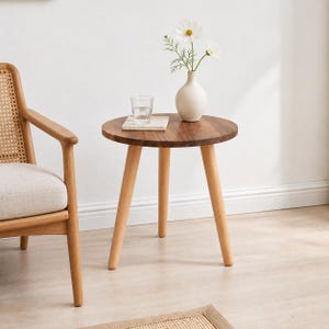 May include: A round wooden side table with three tapered light-colored legs and a dark wood grain top. On the table sits a white ceramic vase containing two white flowers and a clear glass tumbler resting on a small square white coaster.