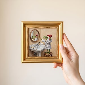May include: A square framed painting featuring a green frog wearing a white bathrobe, standing on a wooden stool in a bathroom. The frog holds a red hair dryer while facing a round wall mirror above a white pedestal sink with toiletries.