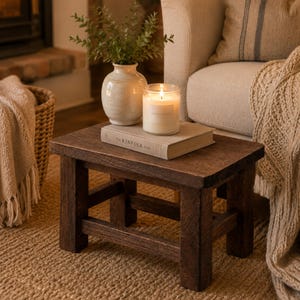 May include: A small, rectangular, dark brown wooden stool with four sturdy legs and horizontal support beams. On top sits a beige ceramic vase containing green foliage, a lit candle in a clear glass jar, and a book titled The Kinfolk Home.