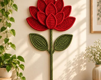 May include: A wall-mounted crochet flower decoration featuring a large, vibrant red bloom with layered, petal-shaped segments. The flower is attached to a long, straight green stem with two symmetrical, pointed green leaves extending outward.
