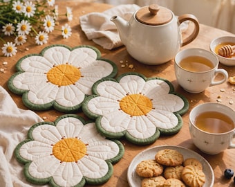 May include: Three quilted fabric coasters shaped like white daisies with yellow centers and green edges. The coasters are arranged on a wooden table alongside a white teapot, two tea cups, a small bowl of honey, and a plate of cookies.