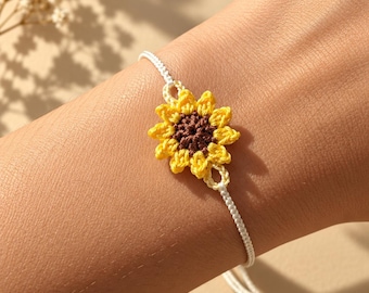 May include: A handmade crochet bracelet featuring a central sunflower motif with a dark brown center and bright yellow petals. The flower is attached to a thin, white braided cord that wraps around the wrist, secured with adjustable sliding knots.