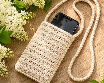 May include: A cream-colored crocheted crossbody phone pouch with a textured stitch pattern and a long matching cord strap. A black smartphone is partially inserted into the top opening of the pouch, which is displayed against a wooden surface.